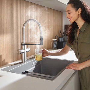 Wasserhahn in einer hellen Küche mit Holzakzenten. Rechts steht eine Frau, die eine Tasse unter den Hahn hält, aus dem kochend heißes Wasser kommt, was man an dem aufsteigenden Dampf erkennt. | Geräte und Ausstattungen in Radevormwald bei MQ Küchen