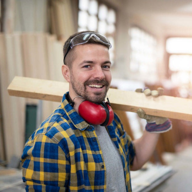 Tischler mit Holz über der Schulter, der freundlich in die Kamera guckt.